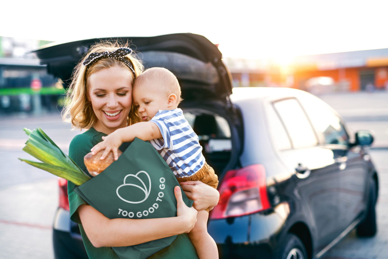 Beautiful young mother with her little baby son in front of a supermarket, holding paper shopping bag. Woman with a boy standing by the car.